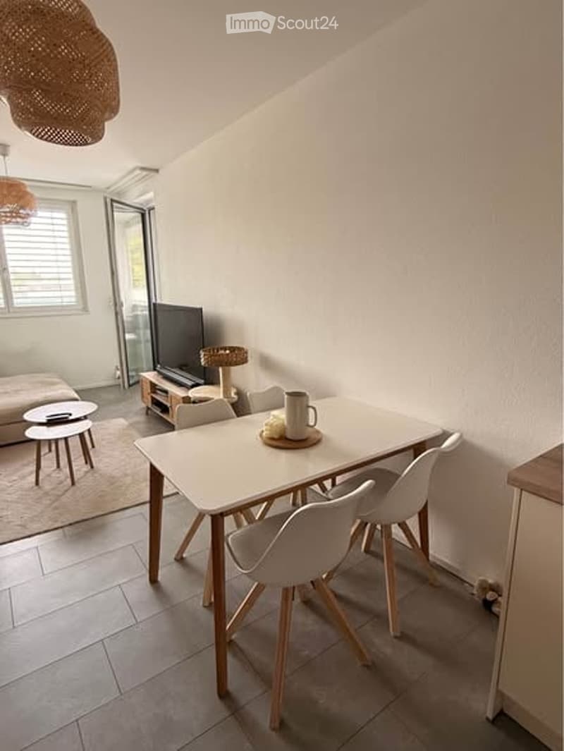 Bright, modern dining and living area with white walls, tiled floor, and large window. Features a white dining table with four chairs, a sofa, coffee table, and a television on a wooden stand.