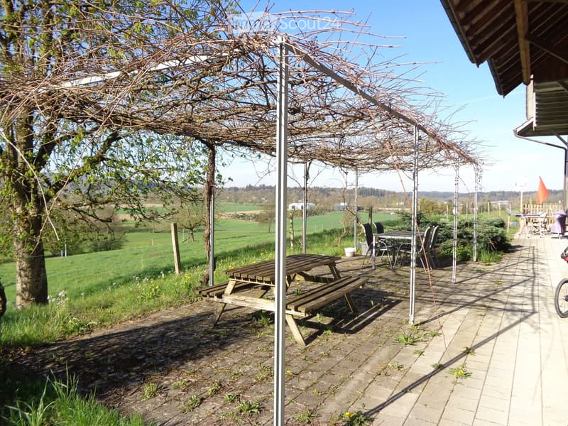 Outdoor seating area with a wooden picnic table, benches, and chairs under a pergola, surrounded by a grassy field and trees.