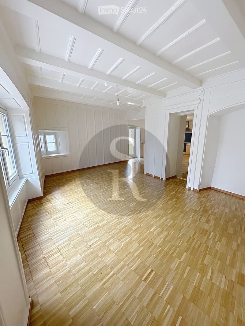 Empty room with light wooden flooring, white walls, and large windows allowing natural light to enter. The space is clean and unfurnished, with visible architectural details such as moldings and a ceiling light fixture.