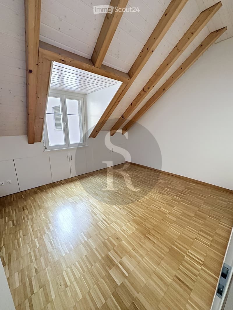 Empty attic room with wooden beams, white walls, and wooden flooring. Features a large window with white frame and a small built-in cabinet.