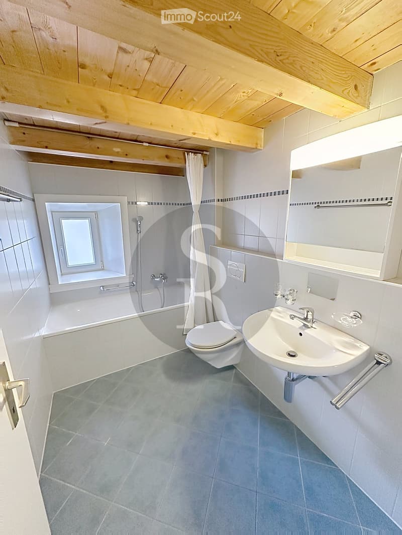 Modern bathroom with a sink, toilet, bathtub, and mirror. Features a wooden slatted ceiling and a window above the bathtub.