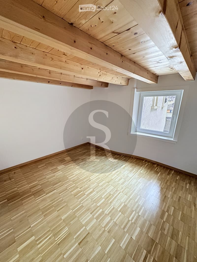 Empty room with wooden flooring and white walls, featuring a single window with a view and exposed wooden beams on the ceiling.