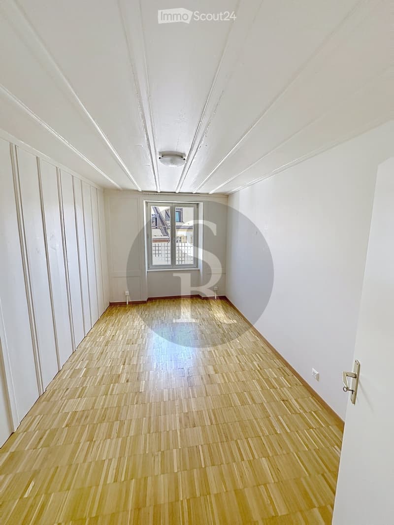 Empty hallway with light wooden flooring, white walls, and a large window at the end.