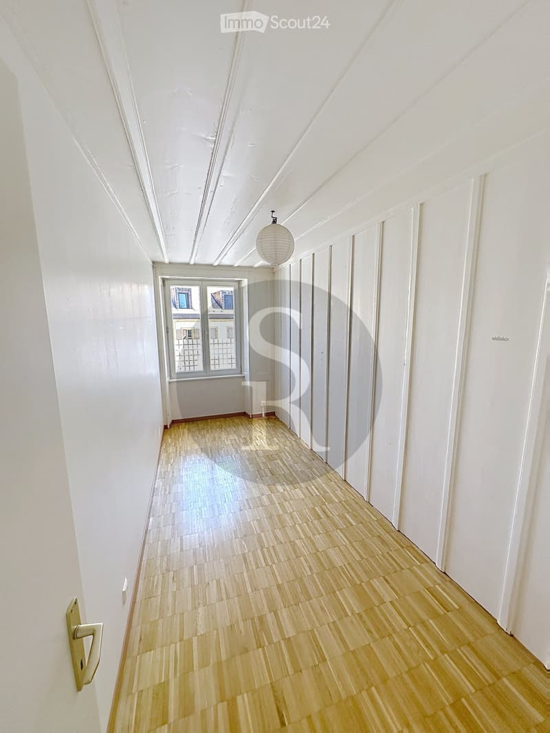 Empty hallway with light wooden flooring, white walls, and a single hanging light fixture. A window at the end provides natural light, and doors line the right side.