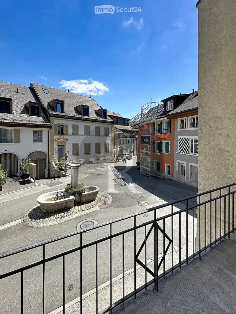 Balcony overlooking a small town square with historic buildings, a fountain, and a few parked cars.
