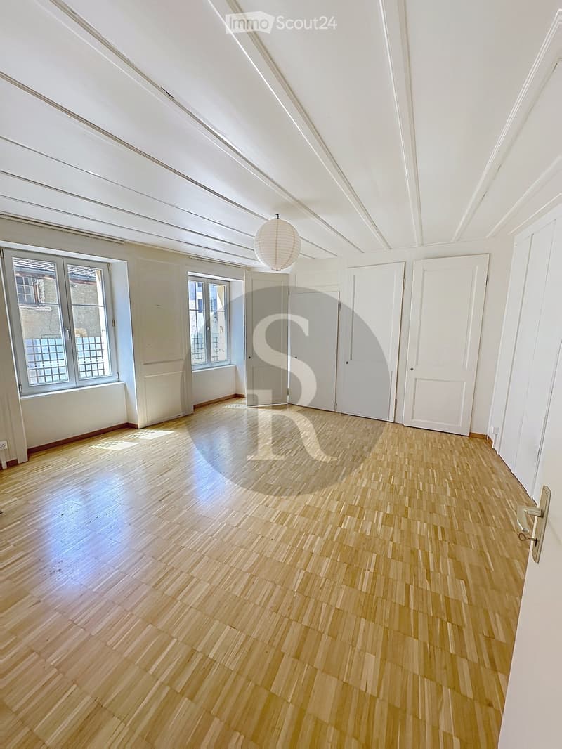 Empty room with wooden flooring, white walls, multiple doors, and large windows allowing natural light. A single hanging light fixture is visible.