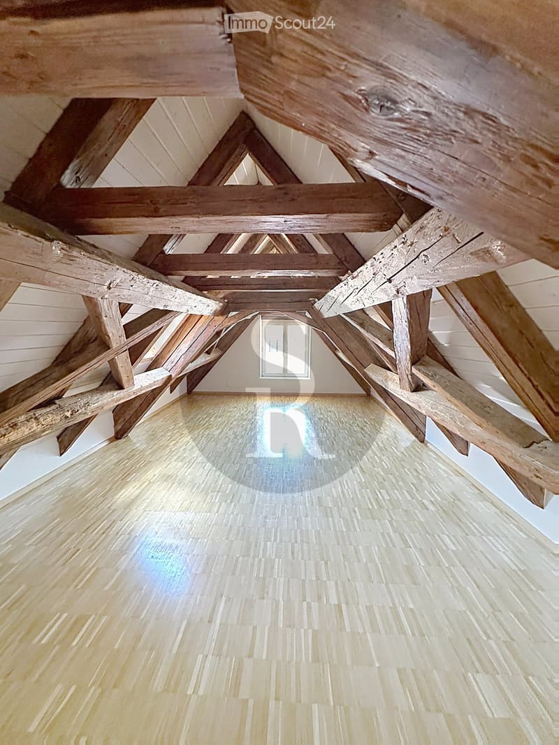An empty attic space with exposed wooden beams and a wooden floor, featuring a small window at the far end.