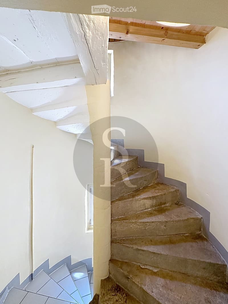 A narrow staircase with beige stone steps and white walls, leading upwards with wooden handrails and a slanted ceiling.
