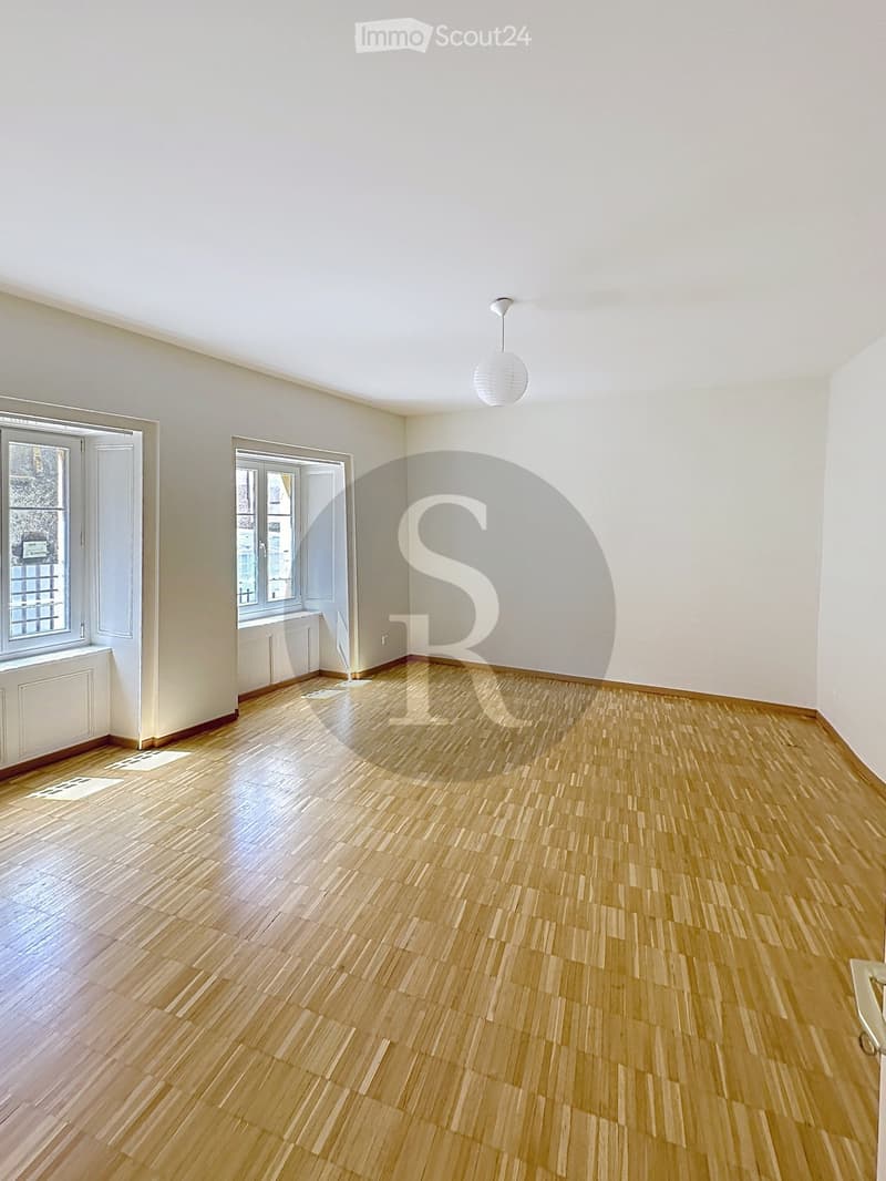 Empty room with light wooden flooring, white walls, and two windows with white frames. A single hanging light fixture is visible.