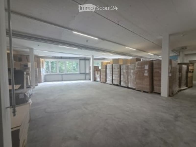 Empty warehouse space with concrete floor, exposed metal beams, large windows, and stacked cardboard boxes along the walls.