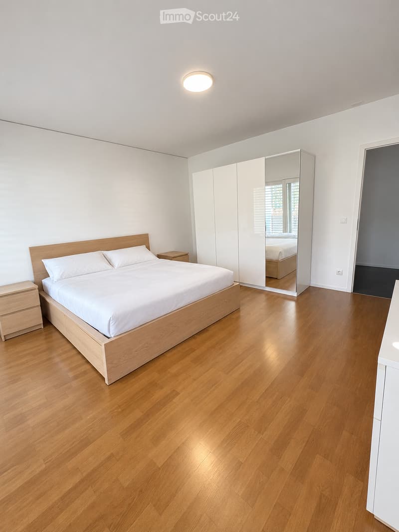 Modern bedroom with a wooden bed, white bedding, nightstands, a large mirrored wardrobe, and hardwood flooring.