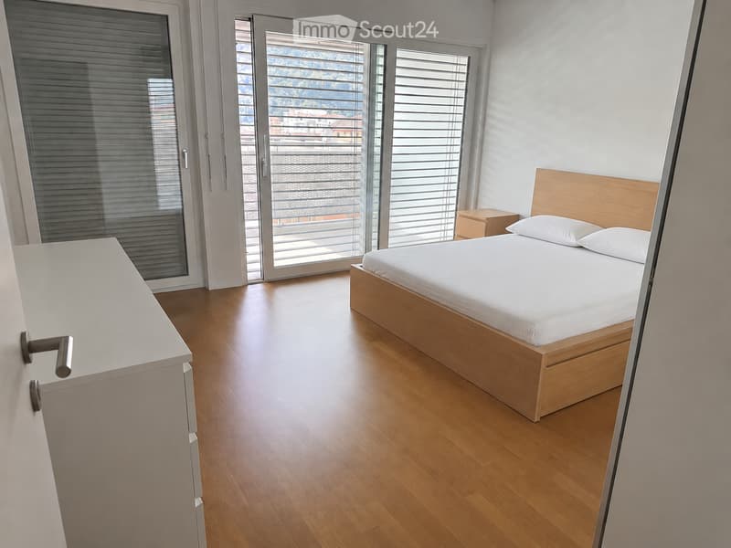 Modern bedroom with wooden flooring, a single bed with white linens, a nightstand, and large windows with blinds offering a city view.