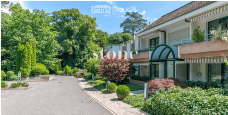 A modern multi-story building with a well-maintained garden and driveway, featuring balconies and large windows.