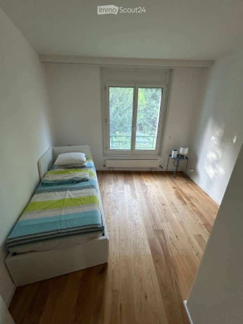 Small bedroom with a single bed, nightstand, and lamp. Light-colored walls and wooden flooring.