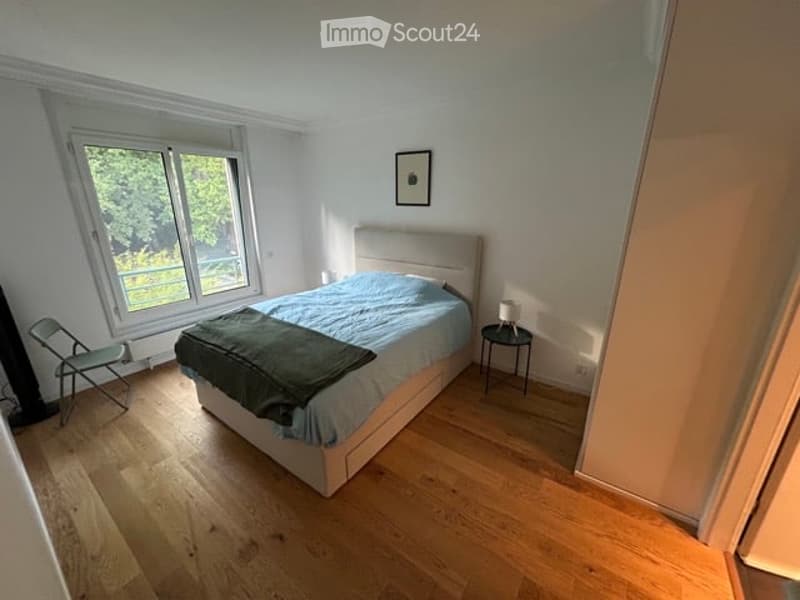 Bright bedroom with wooden flooring, large window, neatly made bed with blue bedding, nightstand with lamp, and a small chair near the window.