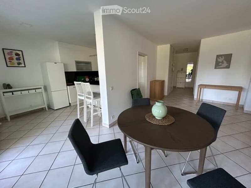 Open-plan living and dining area with a round wooden dining table, four chairs, a refrigerator, microwave, and a small kitchen area with stools.