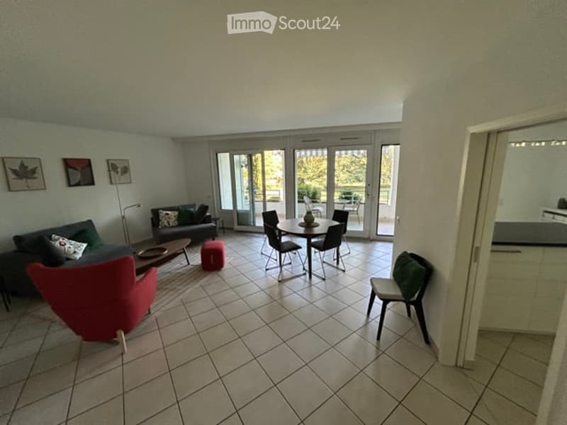 Bright living room with tiled floor, large sliding glass doors leading to a balcony, a red armchair, a sofa, a small table, and a dining area with a round table and chairs.