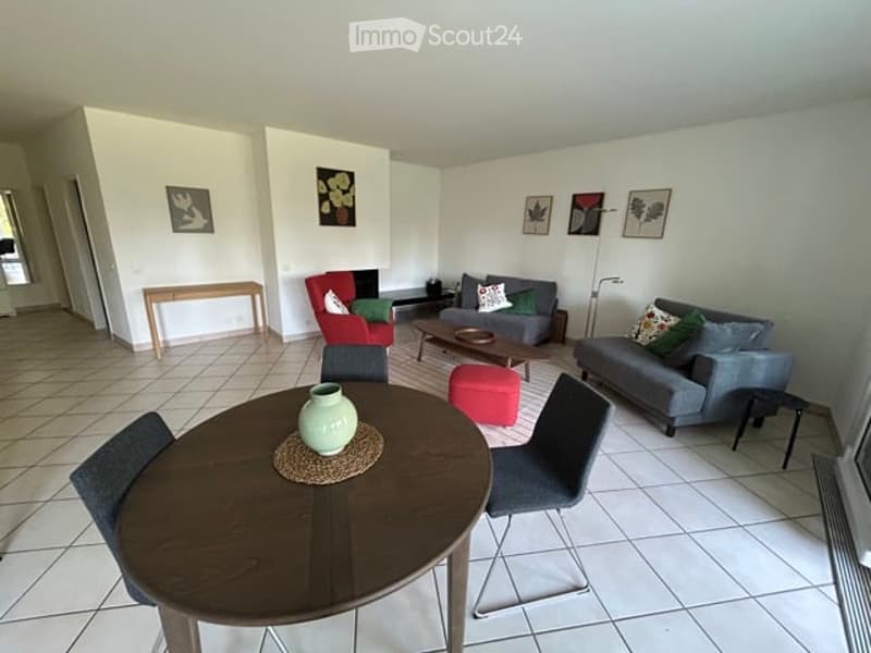 Bright living room with tiled flooring, white walls, and several pieces of furniture including a sofa, coffee table, and dining table with chairs.