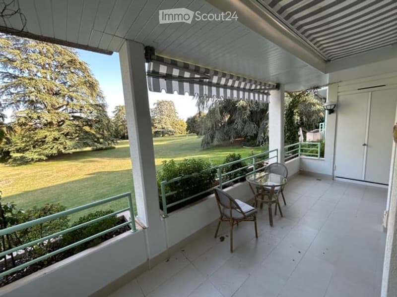 Balcony with white tiled floor, green metal railing, striped awning, small round glass table, and two wicker chairs. Offers a view of a green lawn and trees.