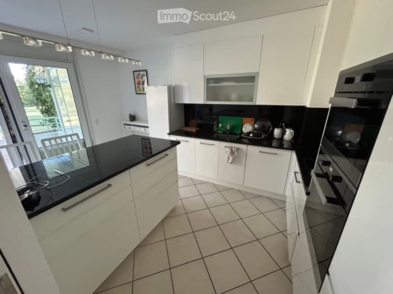 Modern kitchen with white cabinets, black countertops, and tiled flooring. Features include a refrigerator, microwave, oven, dishwasher, and various kitchen items on the counters.