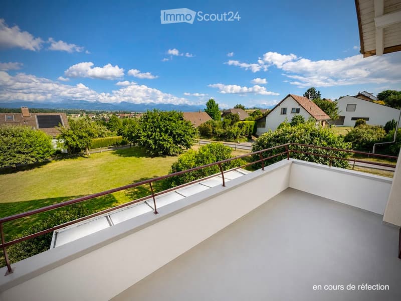 A modern balcony with a white floor and metal railing, overlooking a green lawn, trees, and distant mountains under a clear blue sky.
