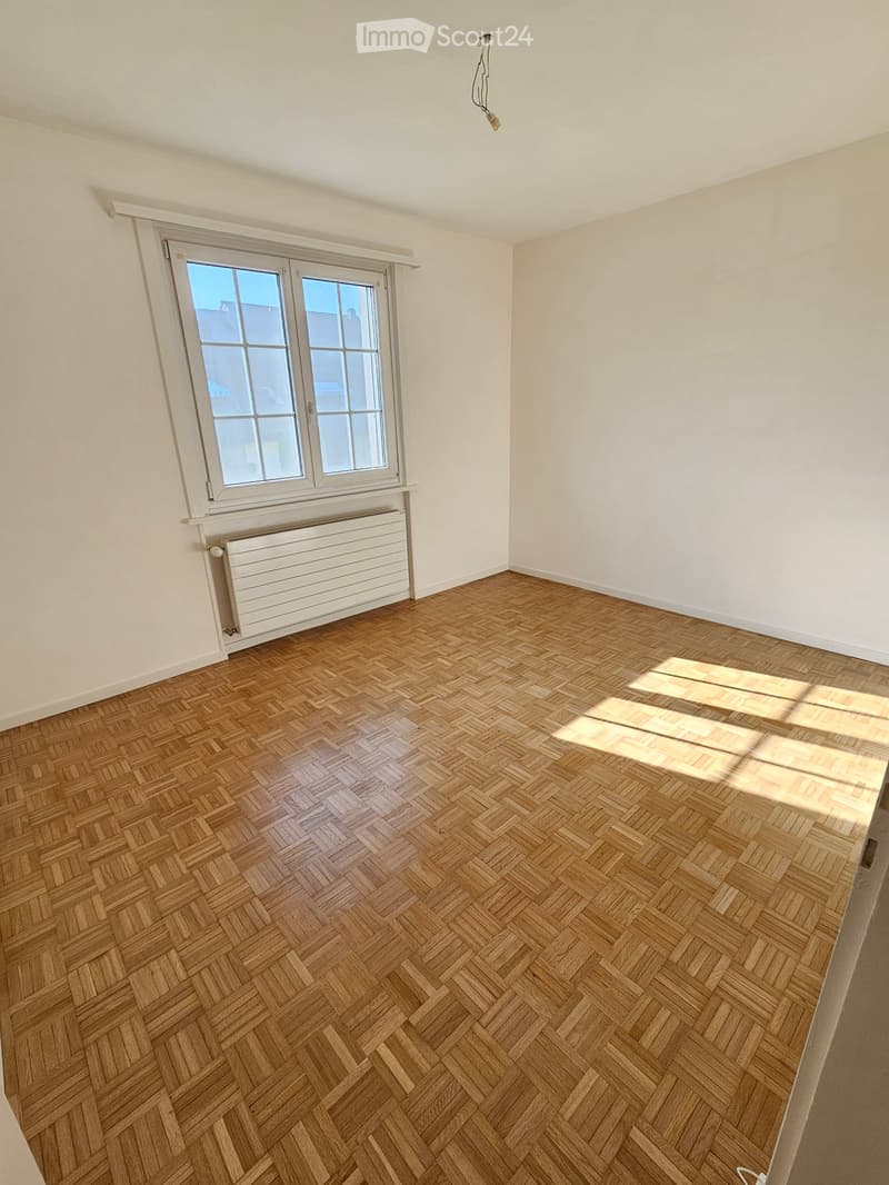 Empty room with wooden herringbone flooring, white walls, and a large window with white frame and radiator below. No furniture or equipment present.