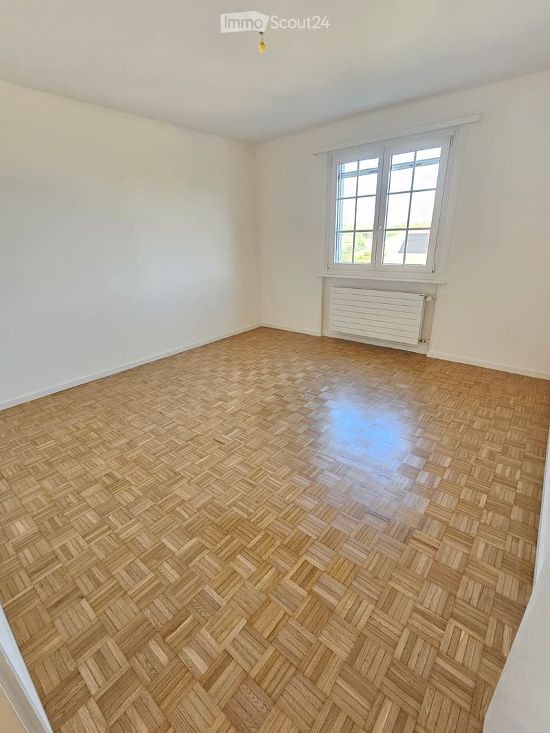 Empty room with light wooden herringbone flooring, white walls, and a large window with white frame and radiator below.