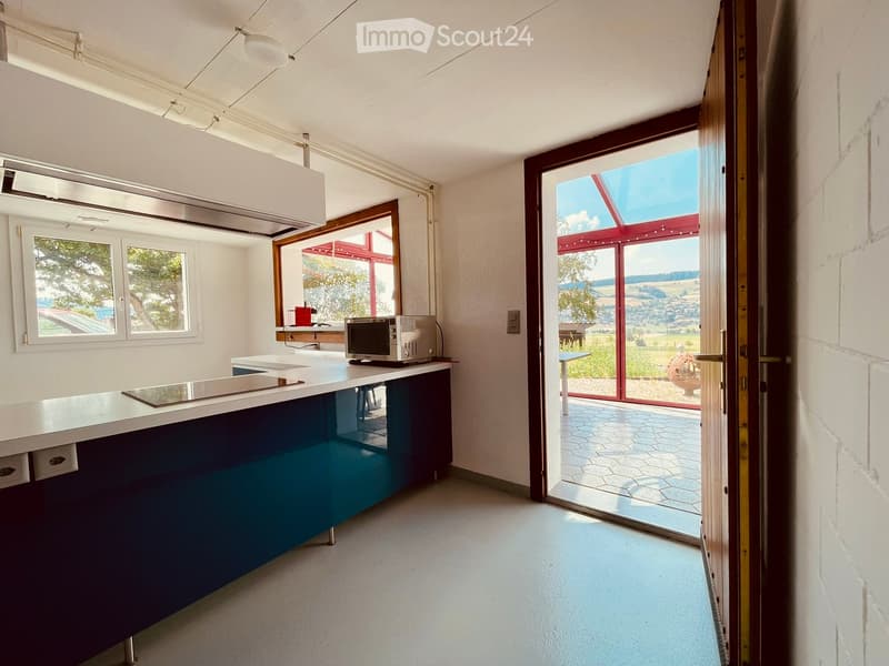 Modern kitchen with white countertops, dark blue lower cabinets, and a microwave on the counter. Large windows and a sliding glass door open to a scenic outdoor view.