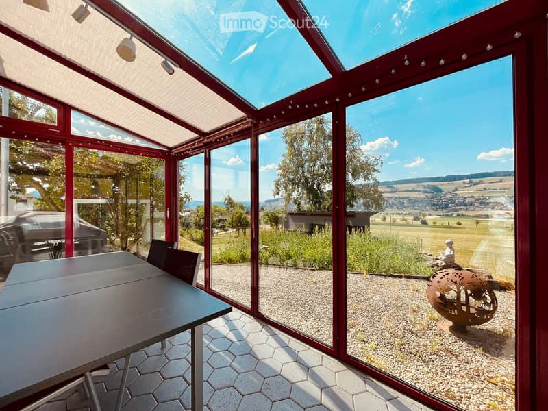 Bright, enclosed patio with large glass walls and a retractable roof, featuring a table and chairs, overlooking a scenic countryside view.
