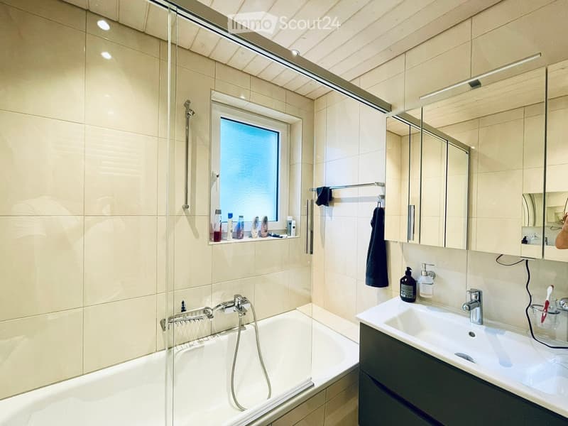 Modern bathroom with a bathtub, sink, mirror, and shower area. Features include a window, tiled walls, and various toiletries on the shelf.
