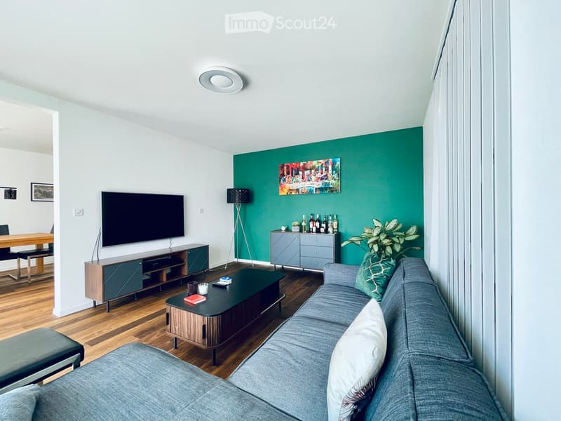 Modern living room with grey sofa, coffee table, TV stand, floor lamp, and a green accent wall with artwork.