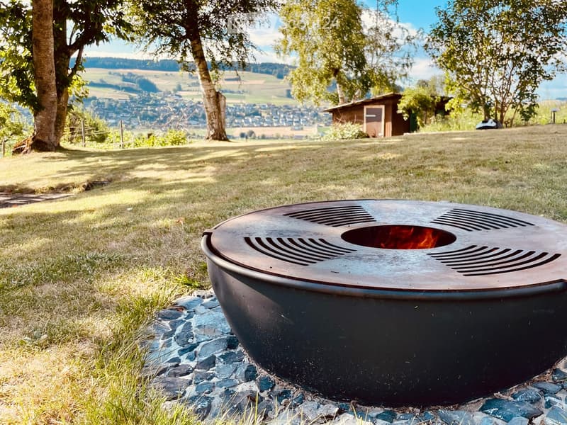A modern outdoor fire pit with a sleek, metallic design is placed on a bed of stones in a grassy area, surrounded by trees and a scenic valley view.