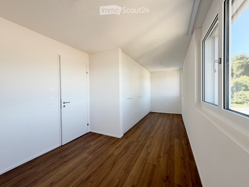 Empty room with white walls, wooden flooring, and large windows offering a view of greenery outside. The room features a white door and built-in cabinetry.