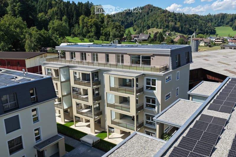 Modern multi-story residential building with solar panels on the roof, surrounded by greenery and a scenic landscape.