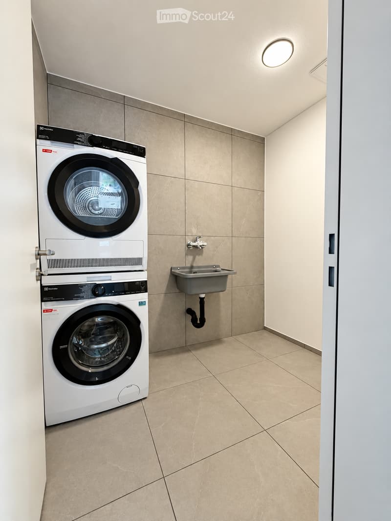 Modern laundry room with stacked washing machine and dryer, a small sink, beige tiled walls and floor, and a round ceiling light.