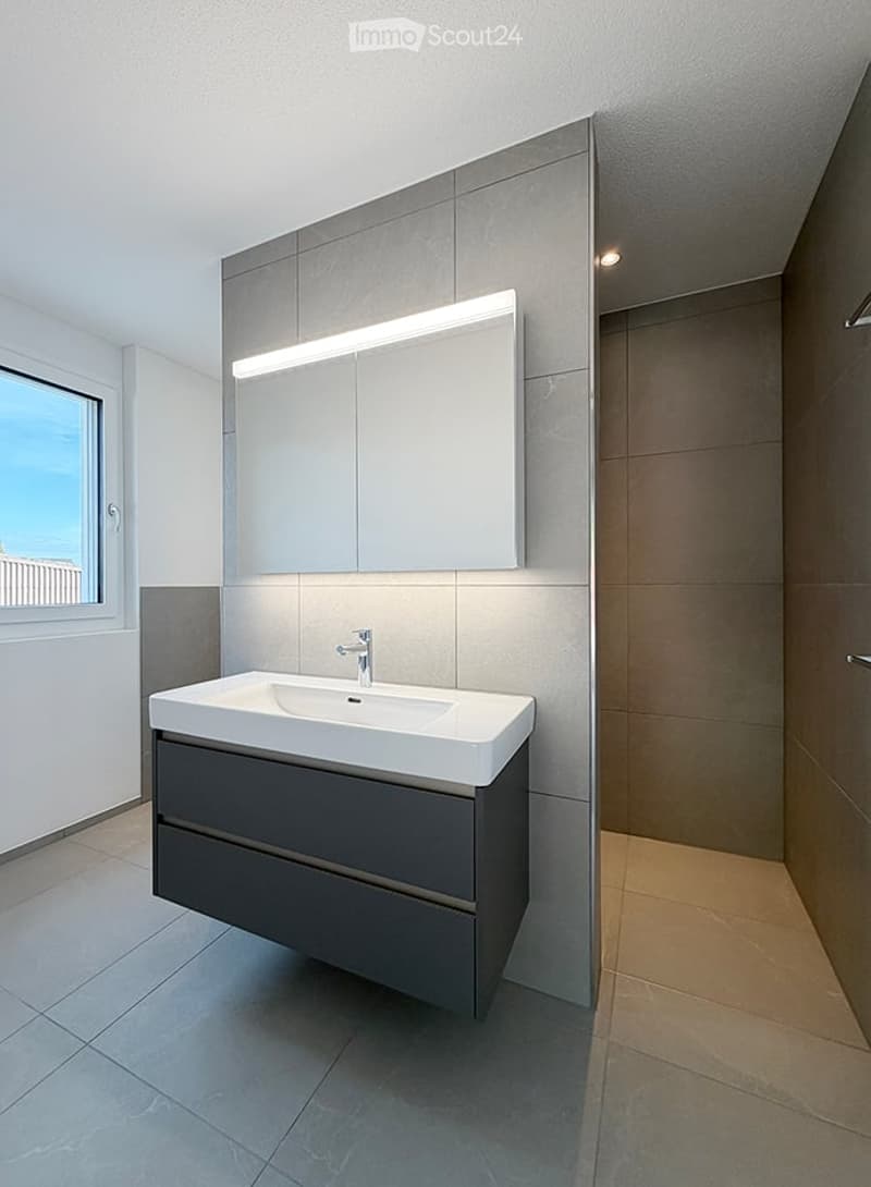 Modern bathroom with a white sink, grey cabinet, large mirror with built-in lighting, and a window offering a view.