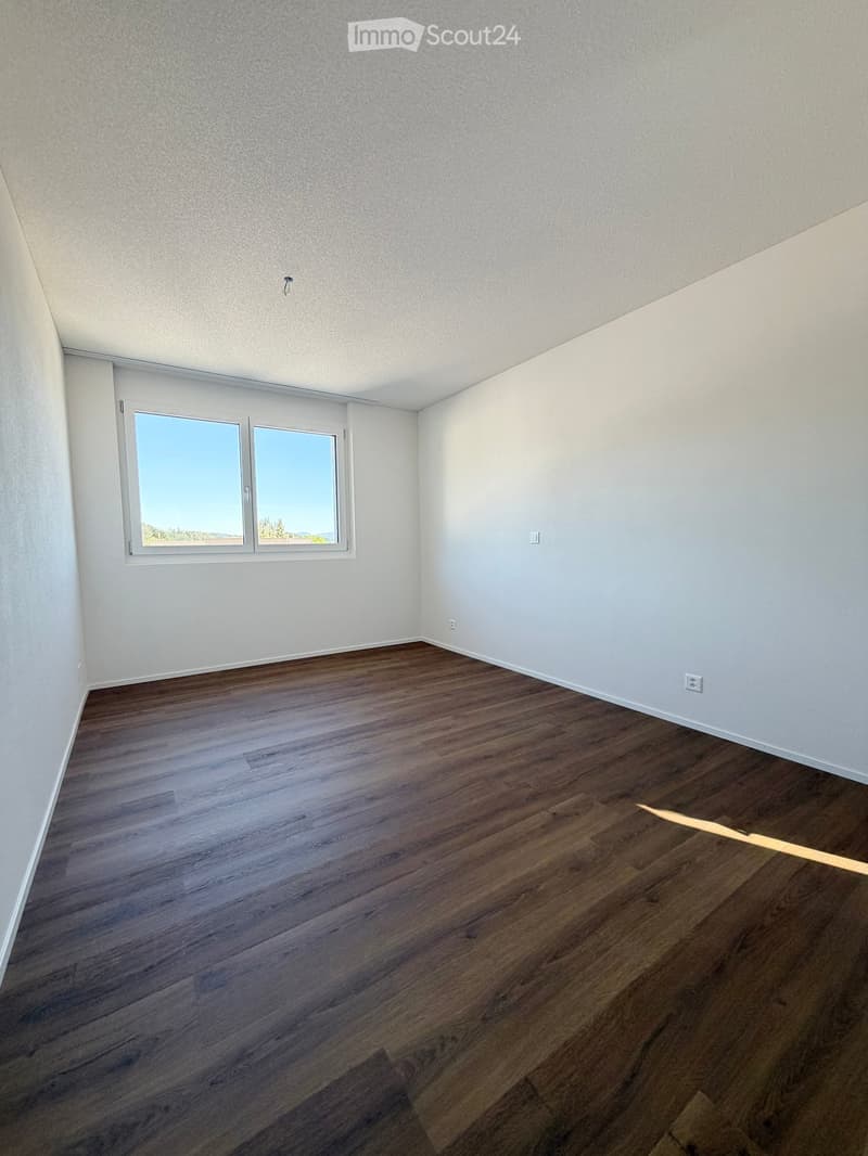 Empty room with wooden flooring, white walls, and large windows offering a scenic view.