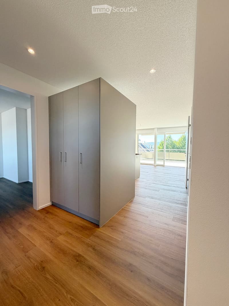 Empty hallway with wooden flooring, white walls, built-in closet, and recessed lighting. The hallway leads to a balcony area with large windows and a view of the outdoors.