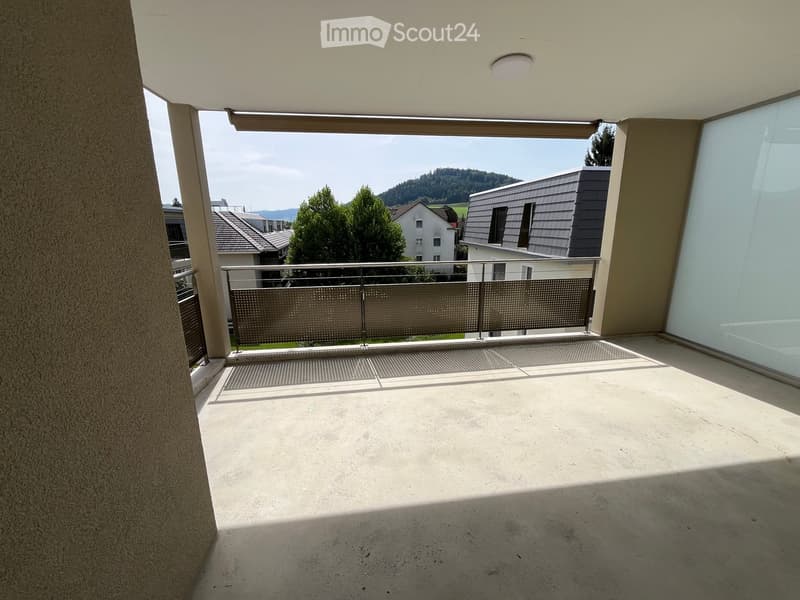 Empty balcony with concrete floor, metal railing, and a view of surrounding houses and greenery.