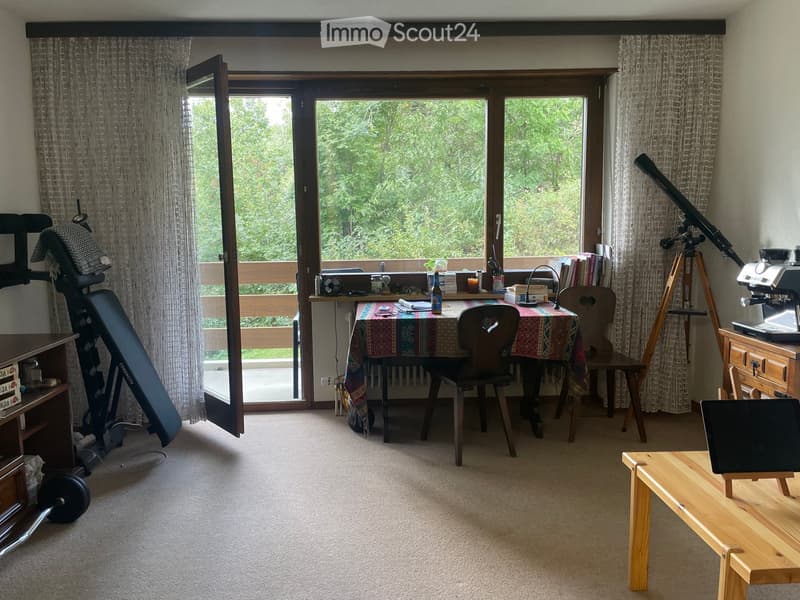 A well-lit room with a large window overlooking greenery, featuring a dining table with chairs, a coffee machine, a telescope on a tripod, and a small wooden bench with a laptop.