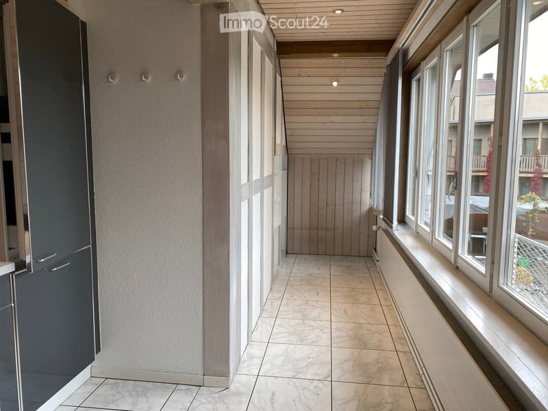 A modern hallway with marble tiled flooring, wooden paneled ceiling, and large windows on one side, leading to a wooden door at the end.