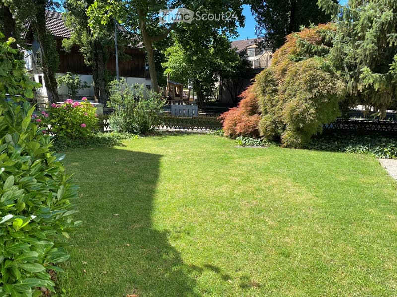 Well-maintained garden with a large, neatly trimmed lawn, surrounded by trees, shrubs, and colorful flowers. A wooden fence and a small play structure are visible in the background.