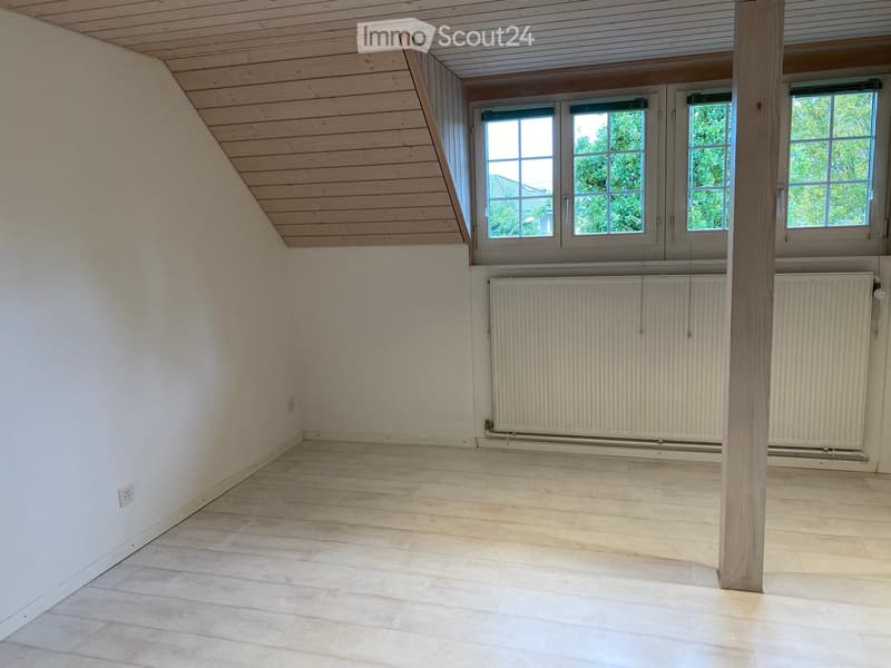 Empty attic room with wooden slanted ceiling, large windows, and radiators.