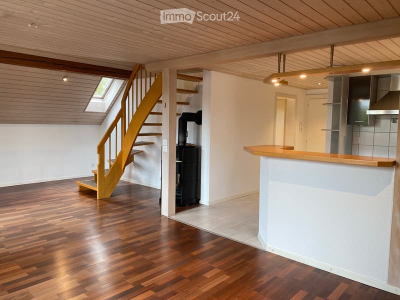 Bright, open room with wooden flooring and exposed wooden beams. Features a staircase leading to an attic, a bar counter, and a wood stove.