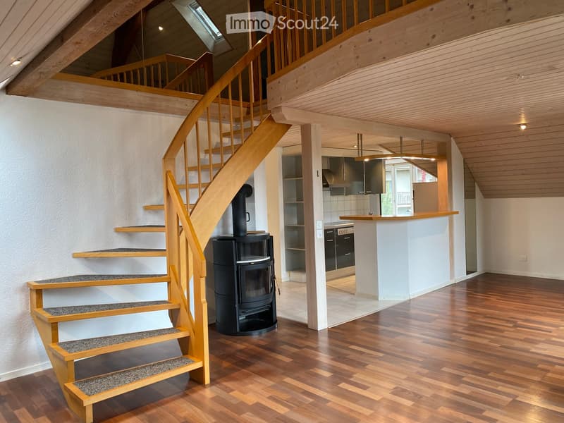The image shows a modern, well-lit interior space with wooden flooring, a wooden staircase with a wooden railing, a wooden ceiling, and a wooden pillar. There is a wooden shelf, a wooden countertop, kitchen cabinets, a kitchen sink, a kitchen stove, and a