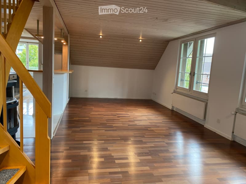 Empty room with wooden flooring, white walls, and large windows letting in natural light. The room features a wooden staircase leading upwards and a small open area with a view of another room.