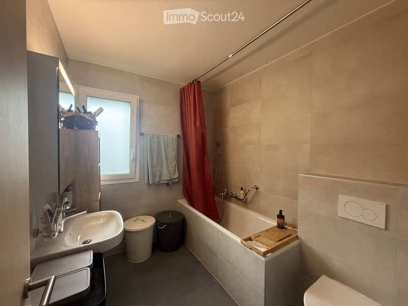Modern bathroom with a sink, bathtub, toilet, mirror, and a window with a red shower curtain. Includes a wooden tray on the edge of the bathtub and two trash bins near the sink.