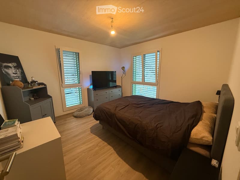 Modern bedroom with wooden flooring, large windows with blinds, a bed with dark bedding, a nightstand, a television, a cabinet, and a chair.