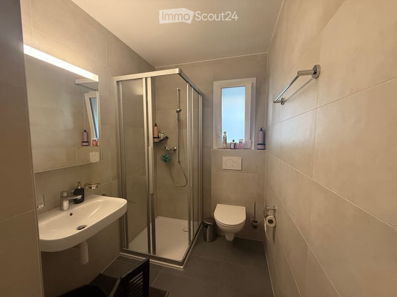 Modern bathroom with a glass-enclosed shower, a sink with a mirror, a toilet, and a towel rack. The space features beige tiles and a window providing natural light.