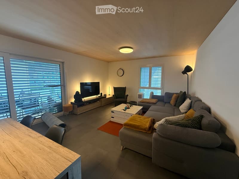 Modern living room with large windows, grey sectional sofa, coffee table, armchair, TV, and a small dining table.